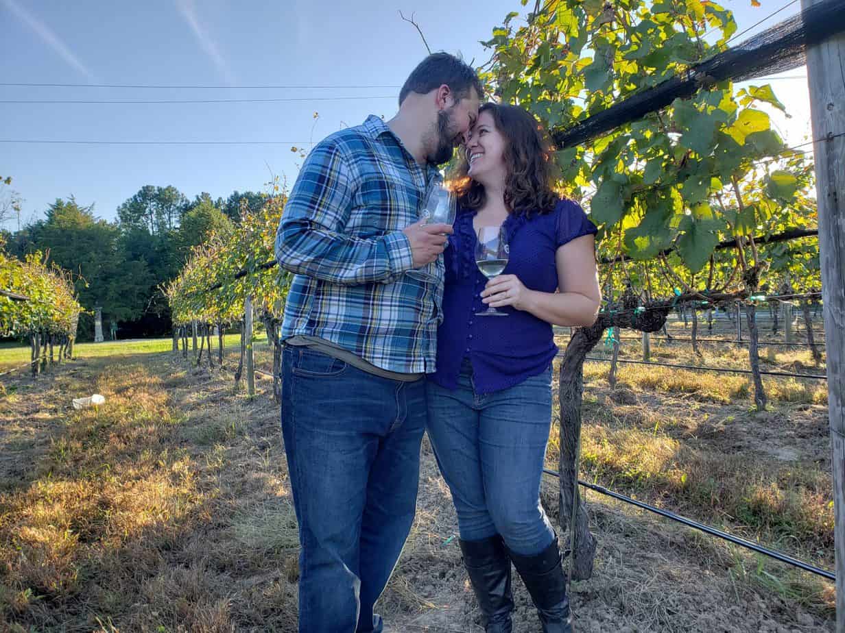 A couple stands forehead to forehead while holding glasses of wine in a vineyard.