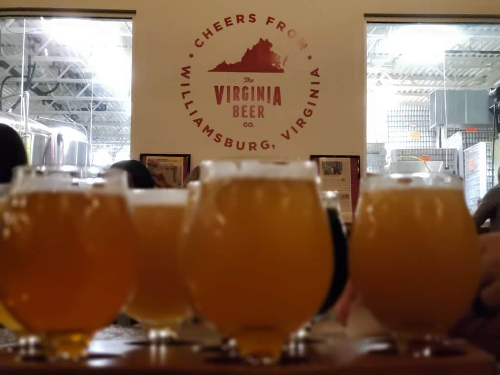 Blurred-out alcoholic drinks sit on a table. Behind, a sign says \"The Virginia Beer Co.\"