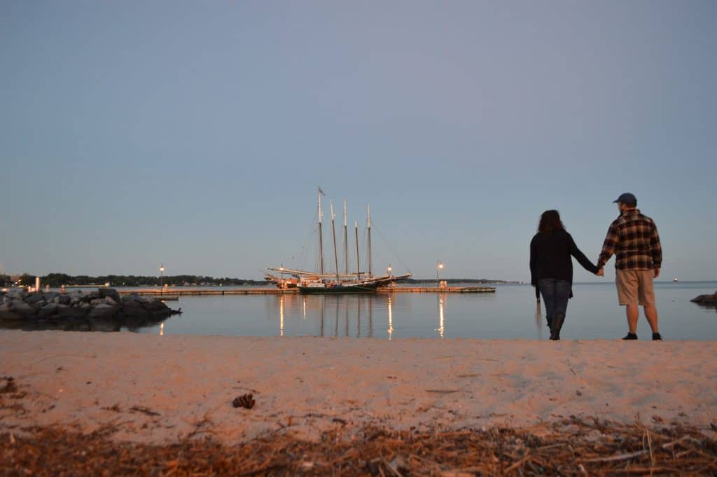 A couple holds hands while looking out at boats docked in the river under a blue sky.