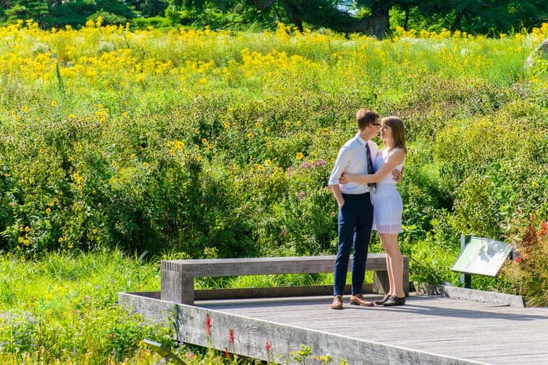 A man and woman smiles on a wooden deck surrounded by greenery.