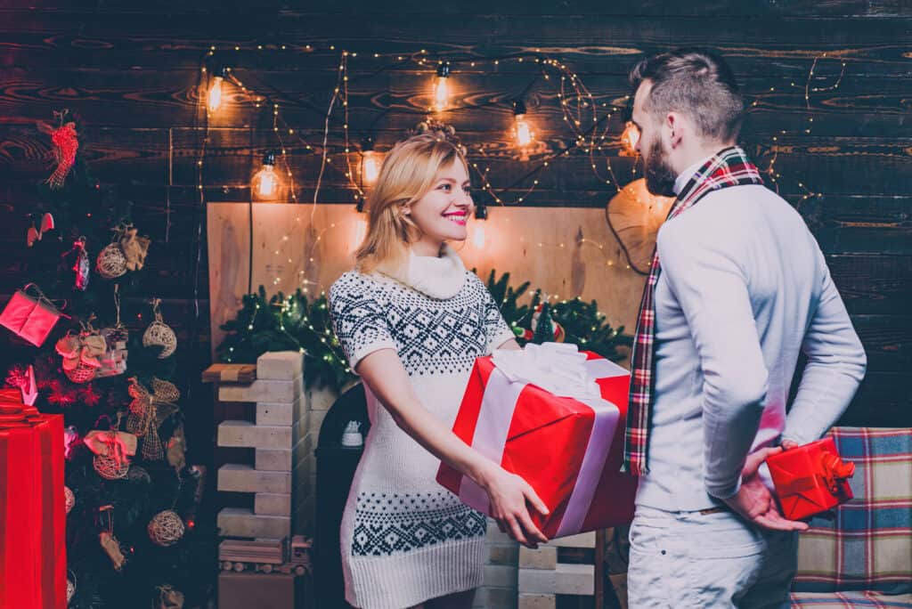 A woman smiles as she hands a big red present to a man in festive holiday clothing.