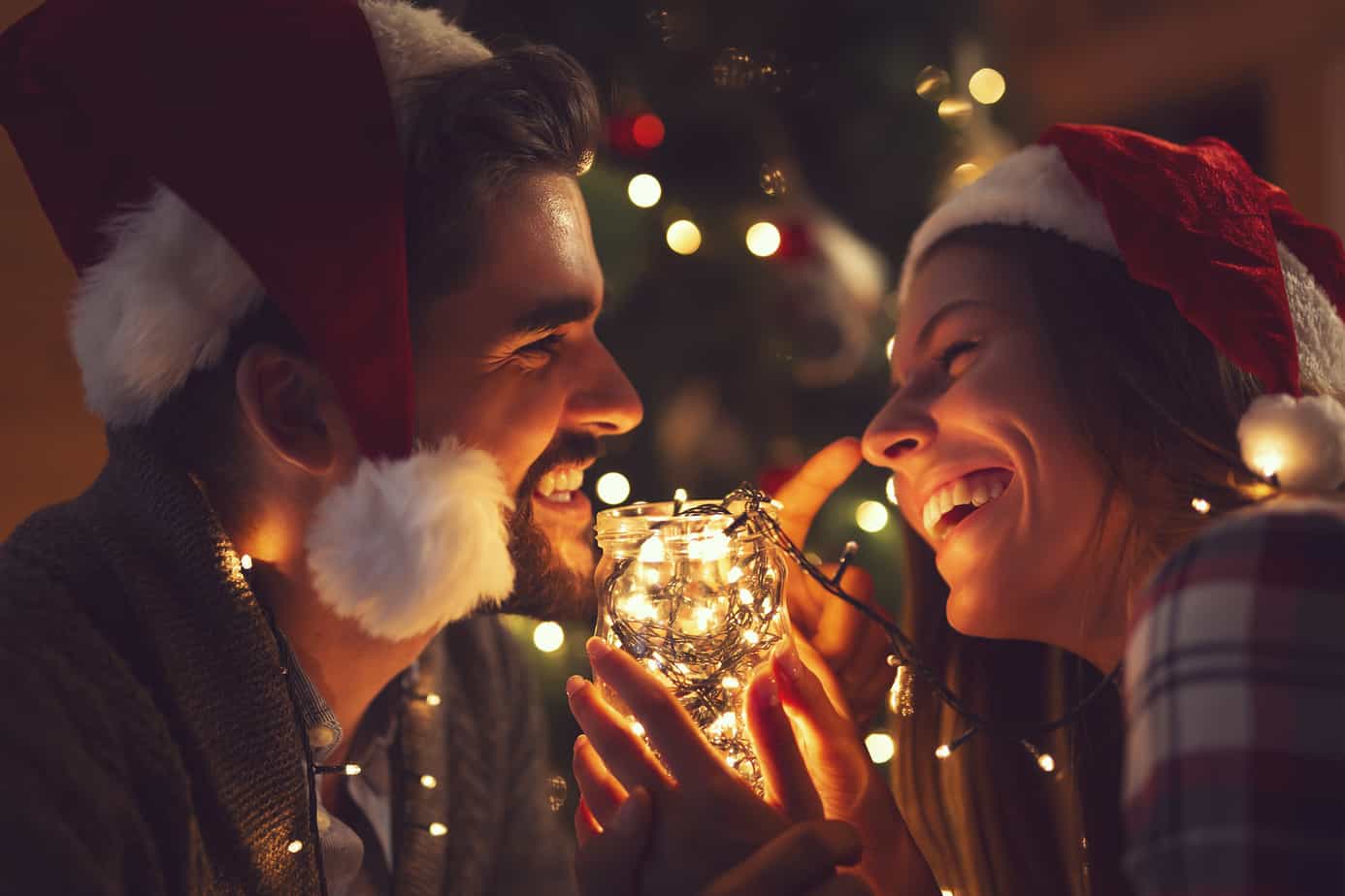 A couple smiles in front of a Christmas tree while wearing Santa hats.