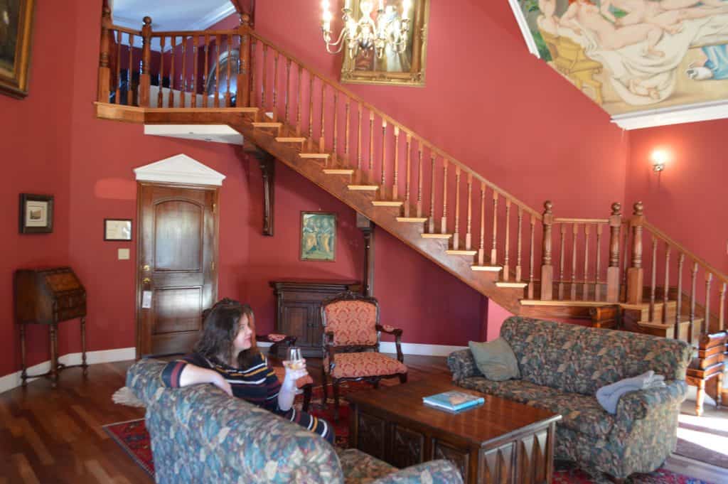 A wooden staircase leads downstairs in a red room. A woman lounges in the sitting area with a wine glass.