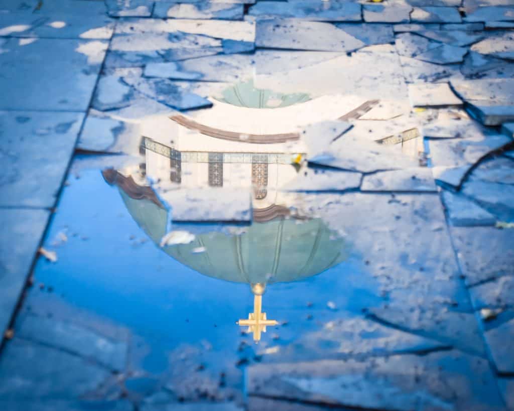 A reflection of a church is seen in a puddle on a sidewalk.