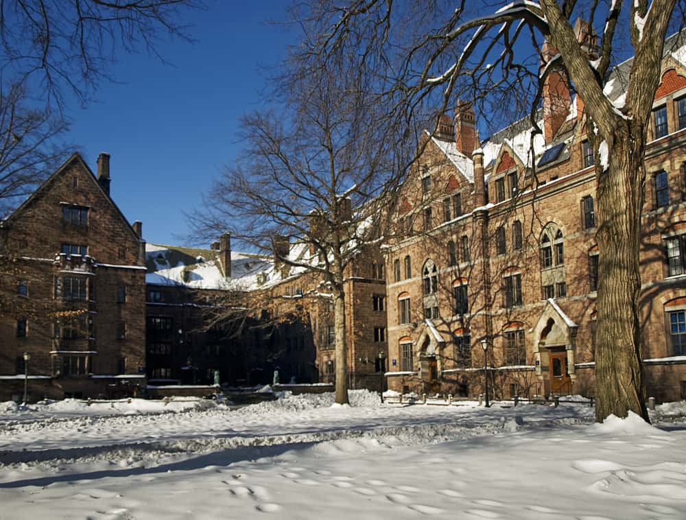 A college campus is covered in snow. The snow in front of the buildings in the background have footprints scattered through them.