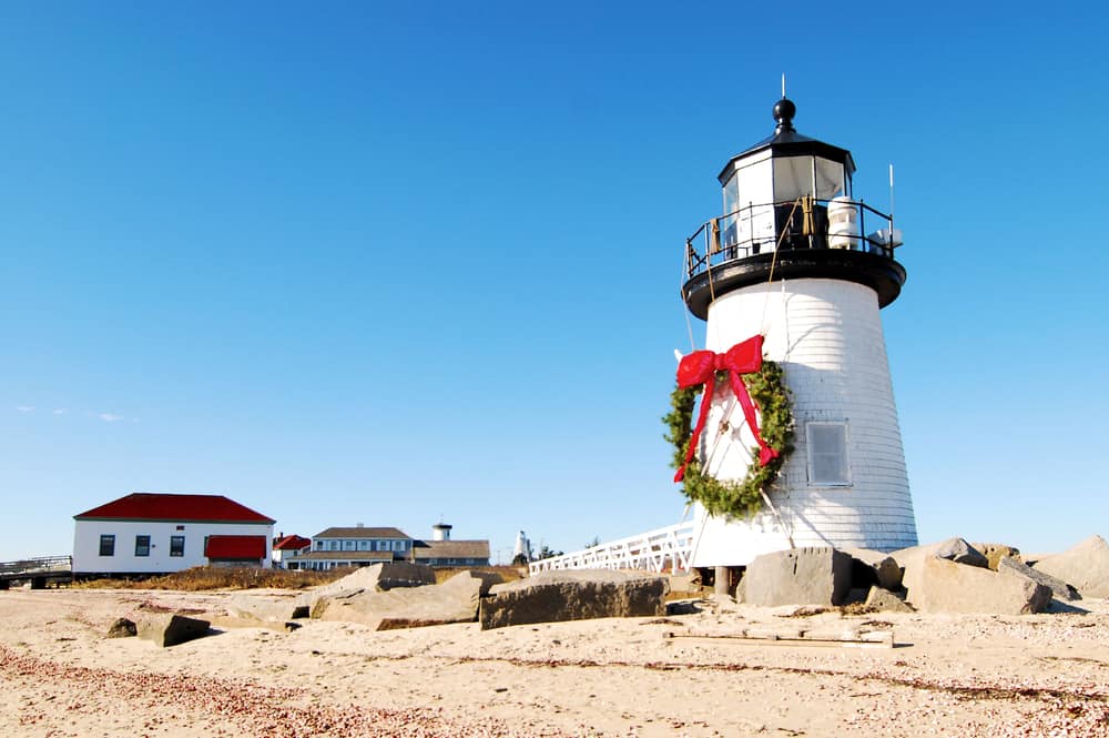A white lighthouse on a beach stands tall with a christmas wreath and big red bow on it.