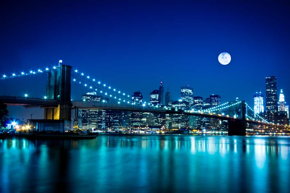A blue-lit photo of a city skyline. A bridge stretches over a body of water.