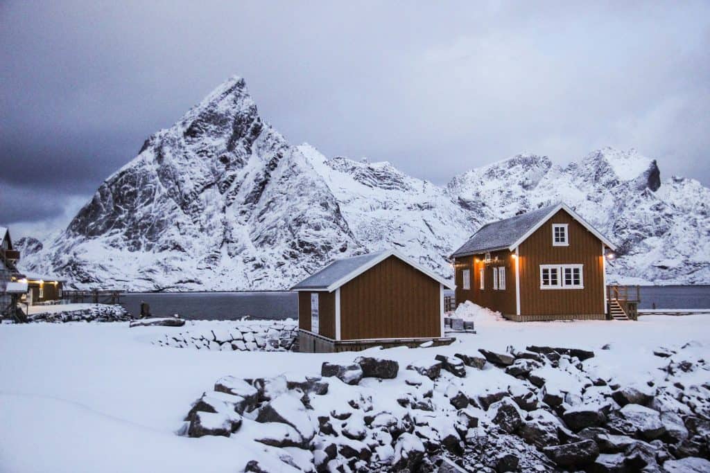 A picturesque snowy village with two buildings in the forefront. A lake is on the other side of the buildings, with tall snowy peaks behind it.