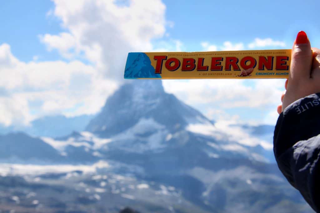 A toblerone bar is held up in hand with a mountain peak behind.