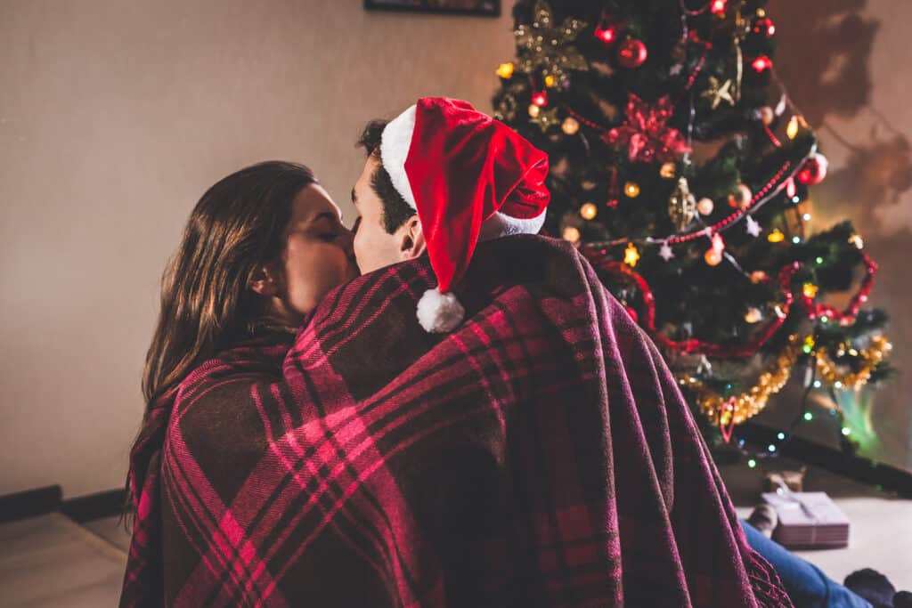 A couple is kissing while wrapped up in a red plaid blanket. The man wears a Santa hat. A Christmas tree is behind them.