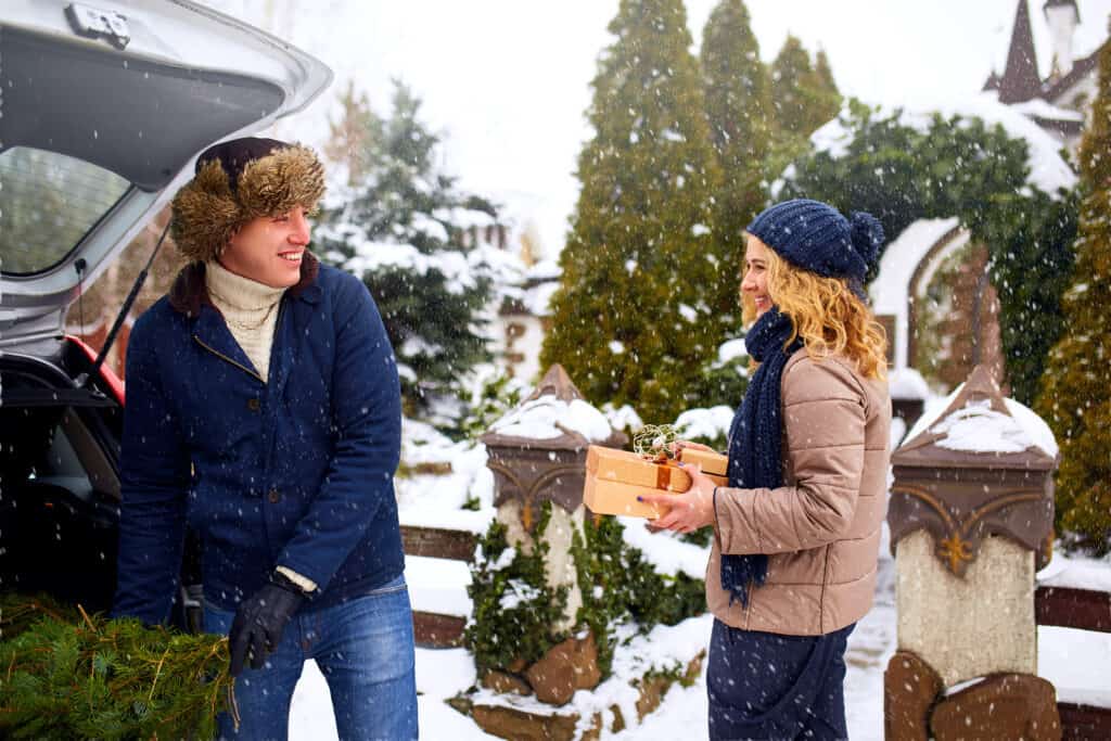 Couple unloading freshly cut down Christmas tree and gift boxes out of car trunk to decorate home. Happy man and woman prepare for New Year holidays together. Snowy winter weather outdoors.