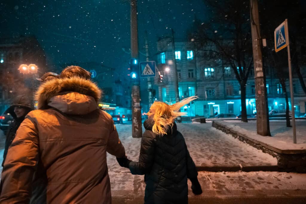 A couple walks outdoors in a city as it snows around them.