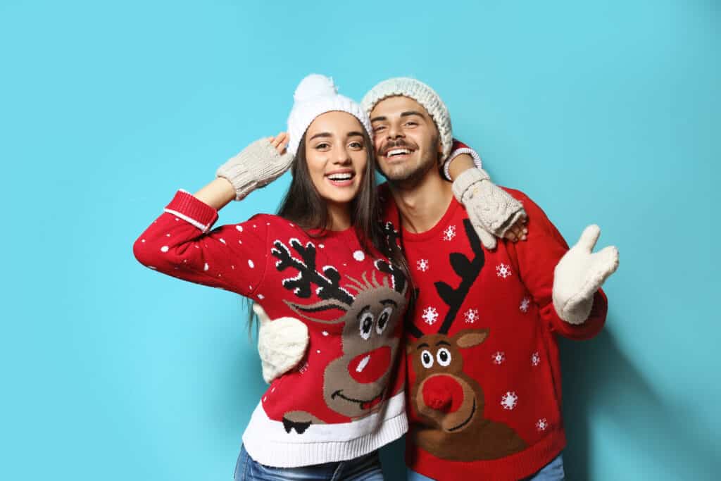 A couple wears Christmas sweaters with reindeer on them in front of a blue wall.