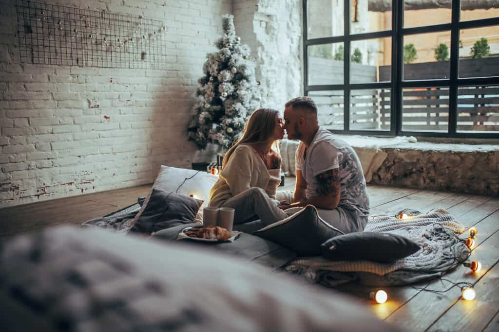 Young couple in love is sitting on floor and kissing next to two cups and plate with cookies on background of Christmas tree, glowing lightbulbs, brick wall and window.