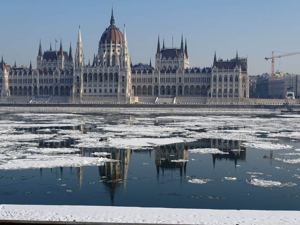 Historic buildings stand tall reflected in a body of water, which is covered in snow.