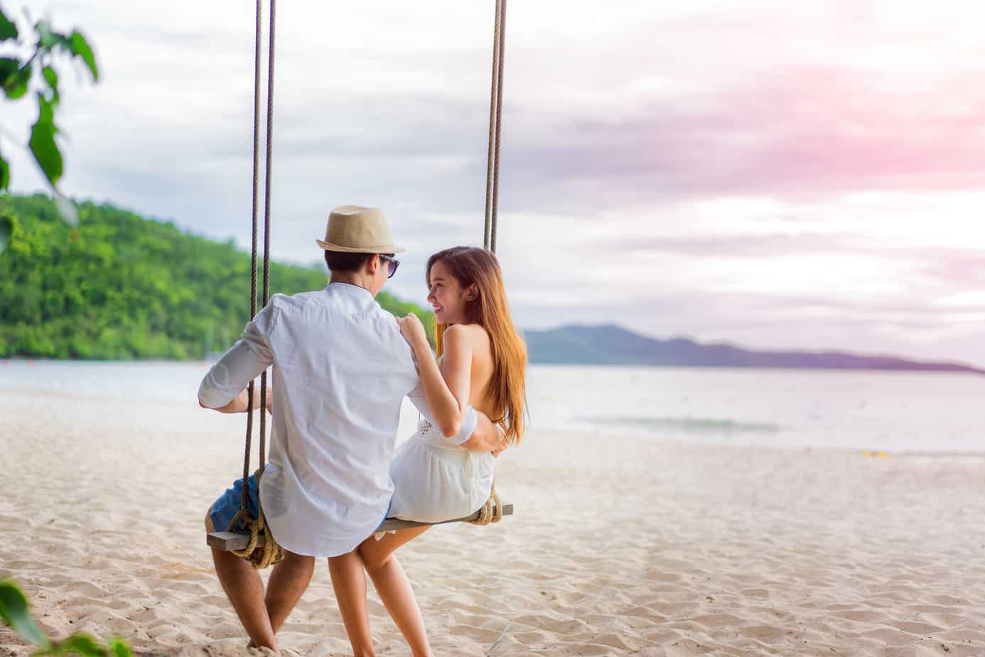 A couple sits on a swing smiling at each other near the ocean.