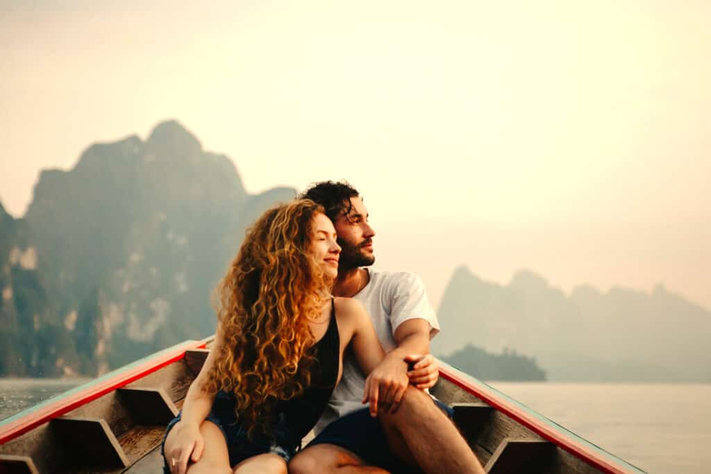 A couple cuddles in a red boat on the water. Mountains can be seen behind them.