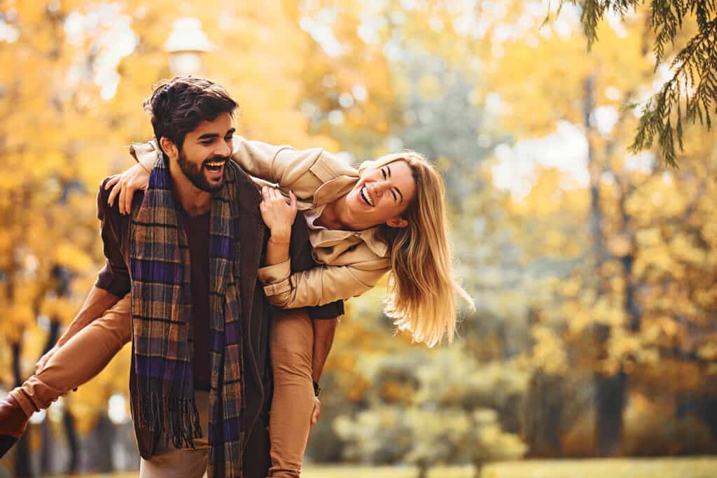 A woman hangs off a man\'s shoulders while they laugh outdoors in the fall.