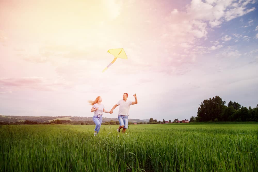 A man and woman run in a field with a yellow kite.