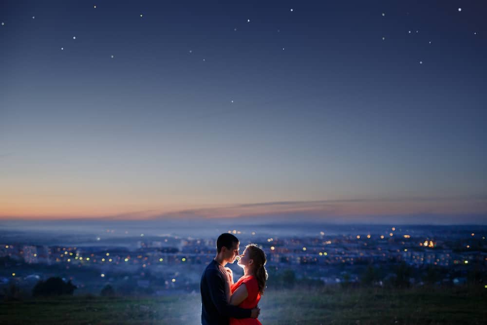 A couple kisses with a town lit up behind them.