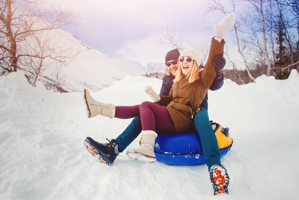 A couple sits on a blue tube smiling in winter gear in the snow.