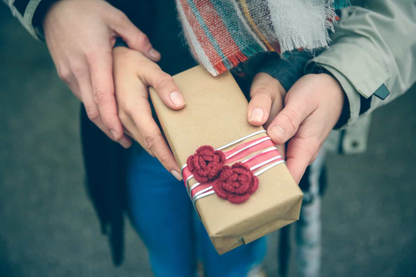 A couple both holds a gift wrapped in parchment paper with yarn flowers.