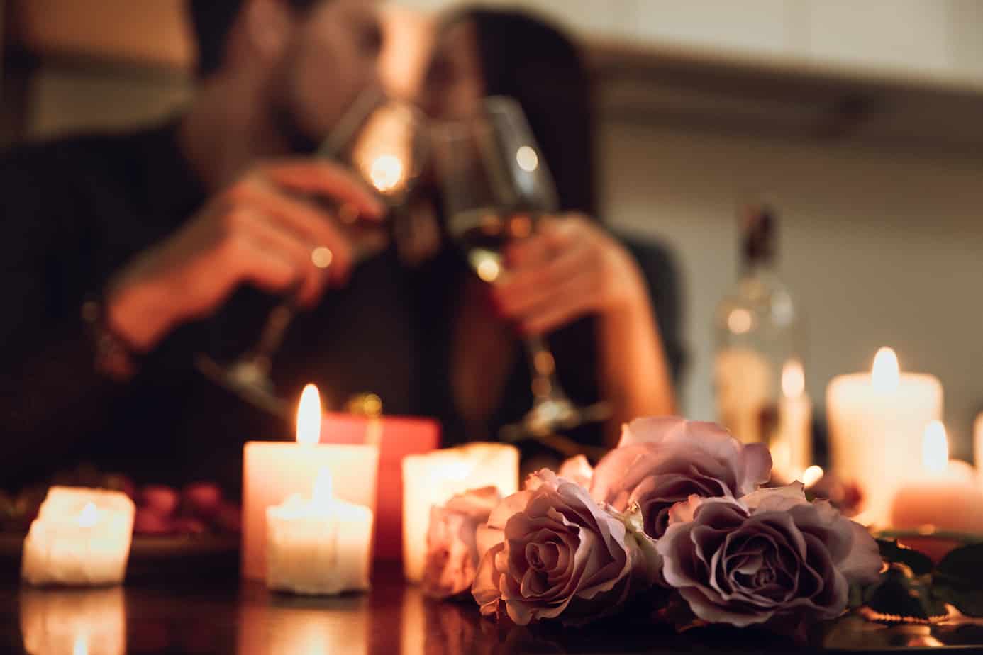 Close up of flowers on a table next to candles. In the background, a couple kisses while clinking glasses.