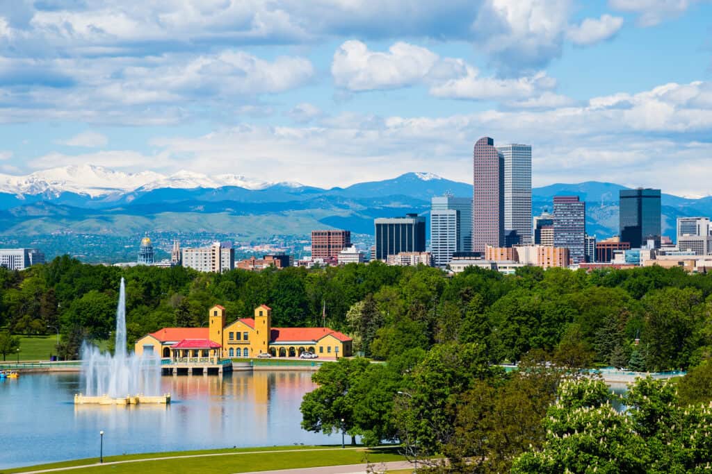 A city skyline with a lake and fountain in the foreground. Snow-capped mountains are in the background.