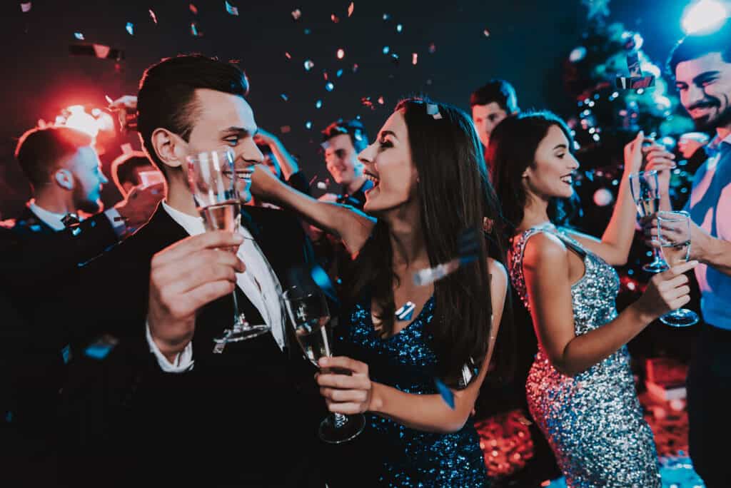 People dance while confetti falls around them. They hold flutes of drinks.