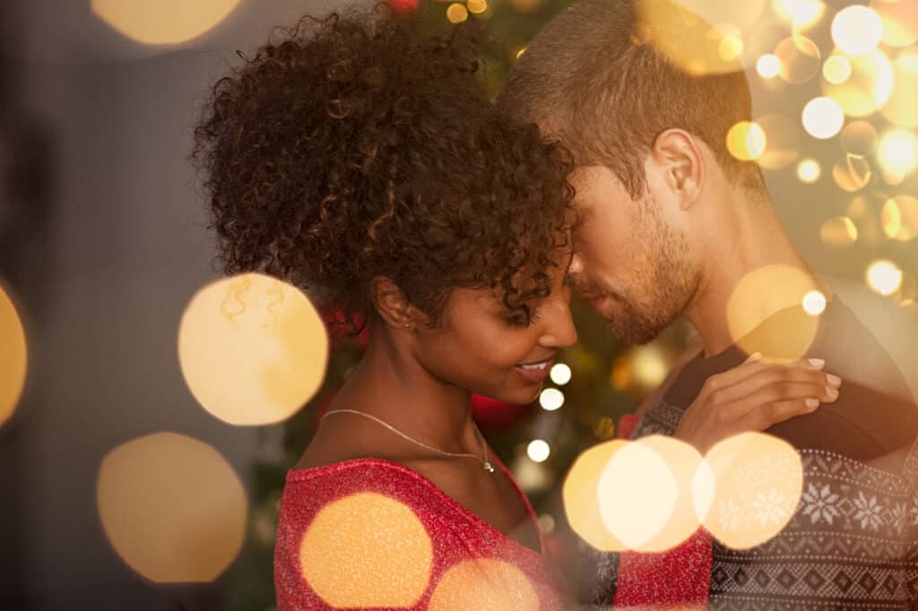 Romantic couple in sweaters dancing before new year eve at home. Young lovely man and african woman hugging over christmas bokeh lights. Multiethnic couple dancing in love with golden lights bokeh.
