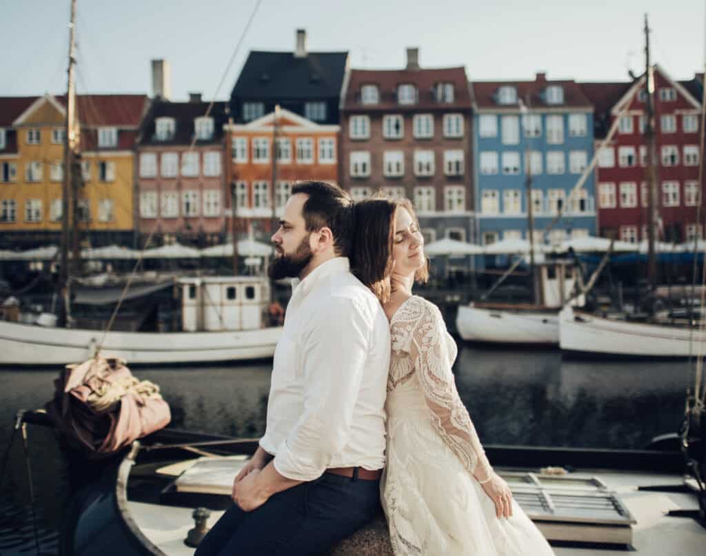 A couple sits back to back with boats in a canal behind them.