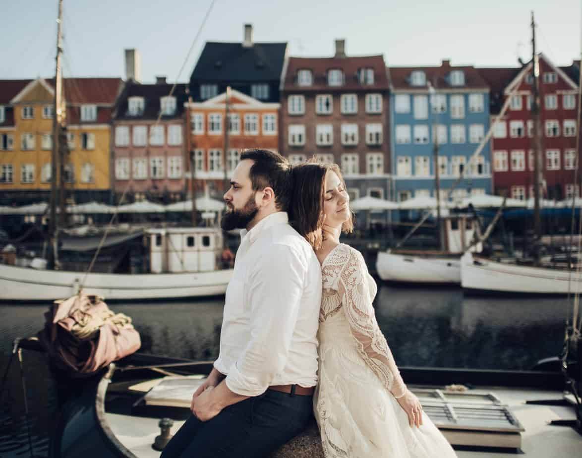 A couple sits back to back with boats with a canal behind them.