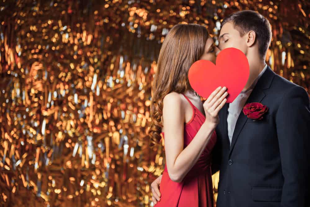 Romantic photo of beautiful couple on glitter gold background. Handsome young man and beautiful woman kissing behind Valentine card in shape of heart