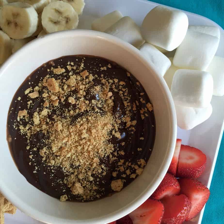 Melted chocolate with graham crackers crushed on top. Around it are marshmallows, bananas, and strawberries.