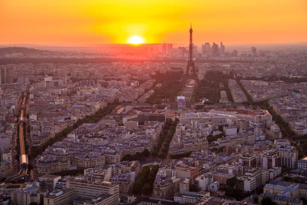 Aerial view of Paris with the Eiffel Tower standing tall. The orange sun sets in the background.
