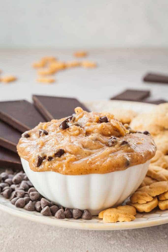Cookie dough overflows out of a white bowl surrounded by chocolate and nuts.