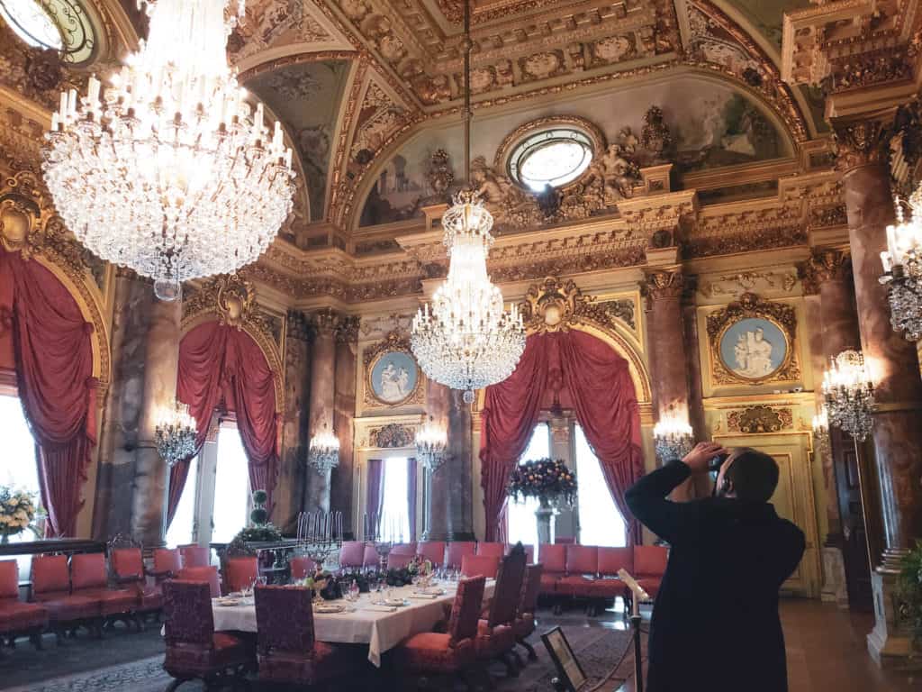 A man takes a photo of a chandelier hanging down from a ceiling in an over-the-top dining room.