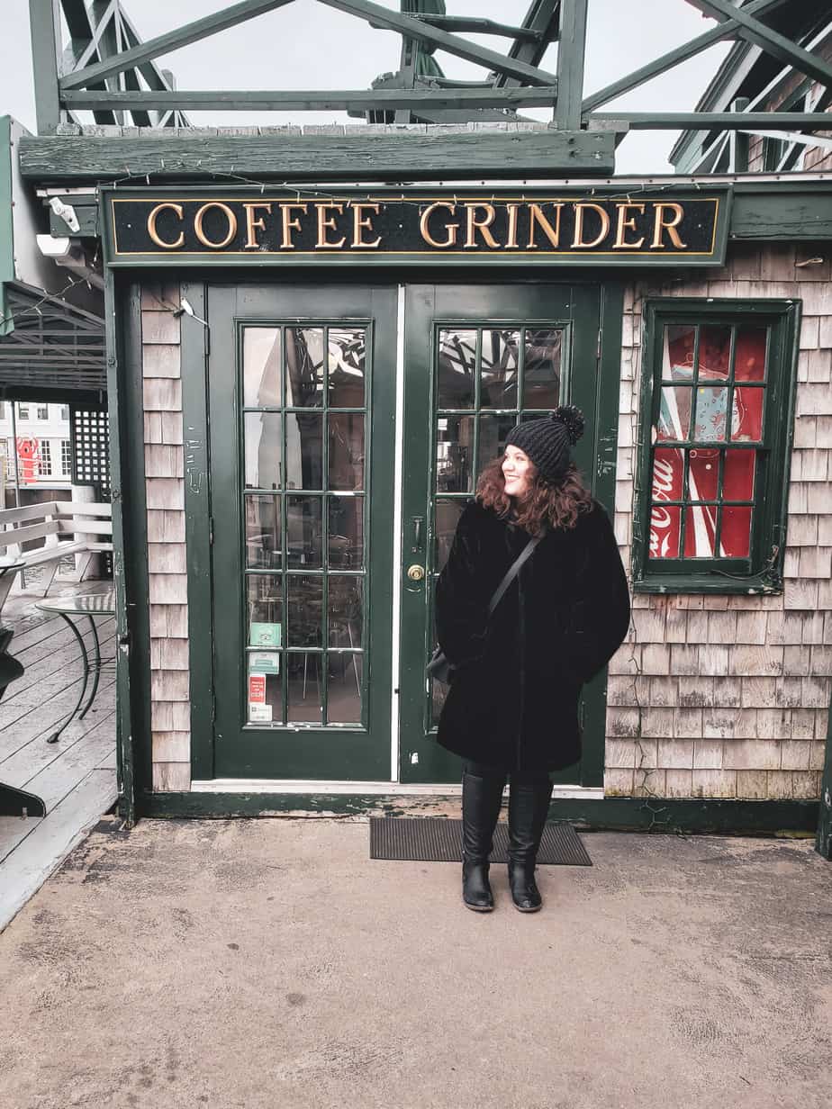 A woman stands all bundled up in front of a cute shop that says Coffee Grinder.