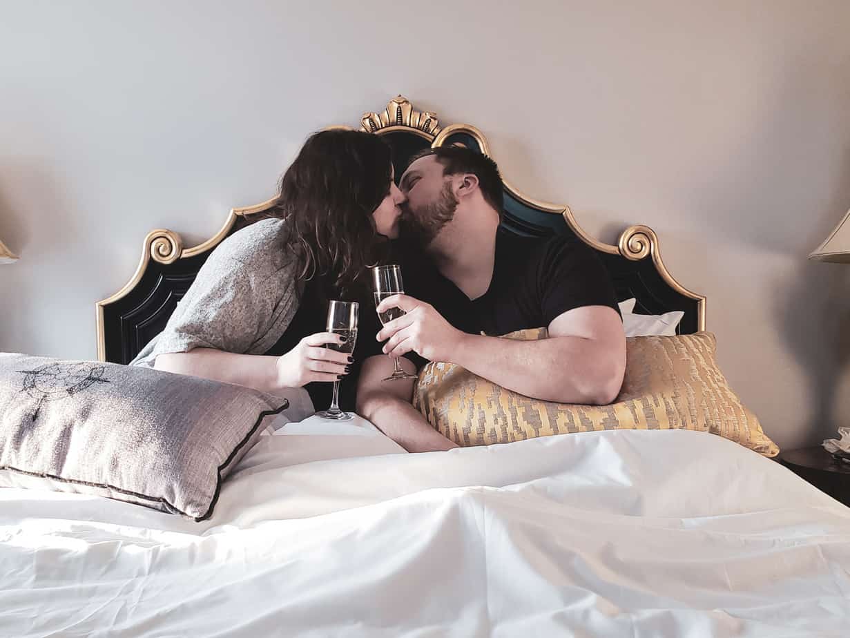 A couple kisses while holding wine glasses in a bed.