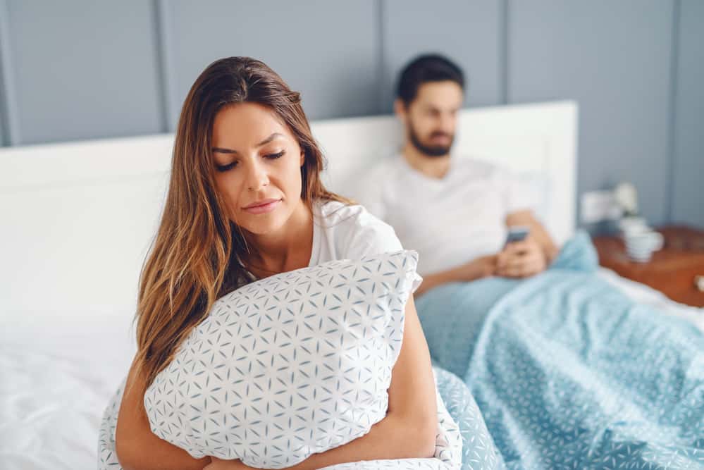 A woman hugs a pillow while a man is on his phone. They both look upset.