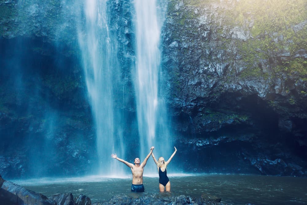A couple stands holding arms up with a waterfall behind them.