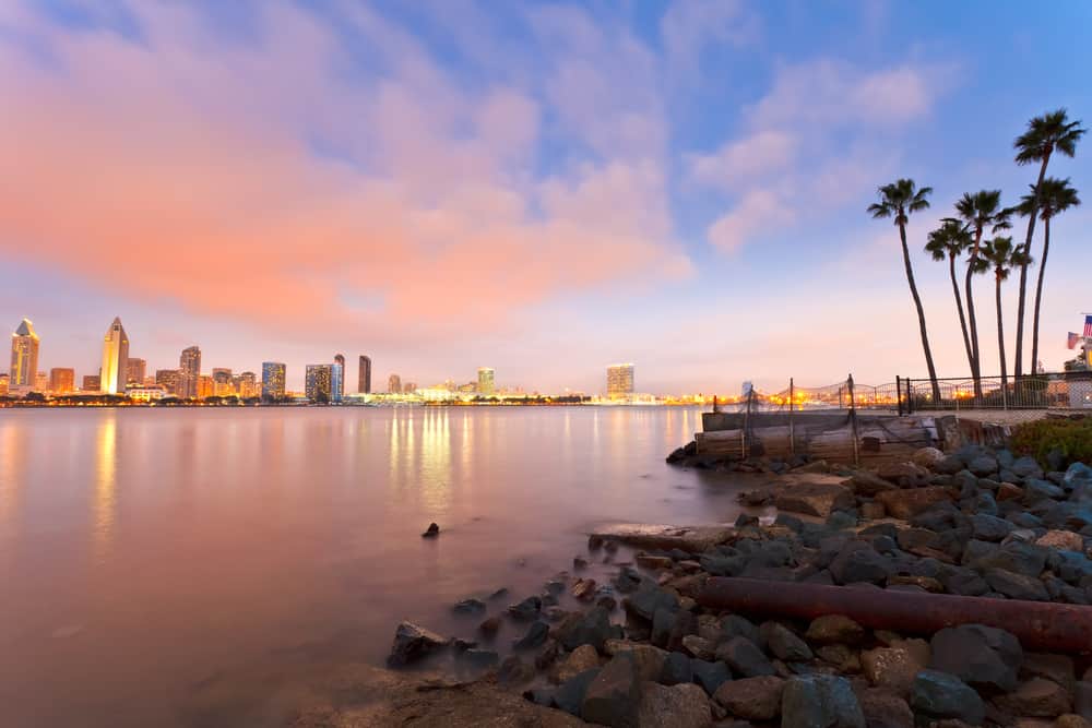 A sunset over a hazy body of water. In the foreground are rocks and palm trees. The background has a city skyline.