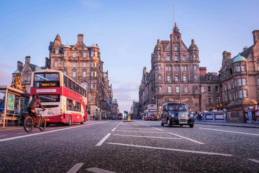 Double-decker buses and other cars driving on opposite sides of the road in between large European styled buildings. 