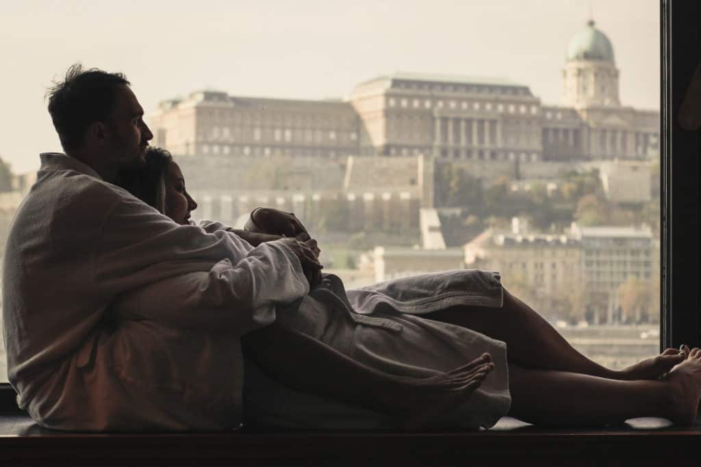 A couple snuggles together in robes at a window looking out on a romantic European honeymoon destination