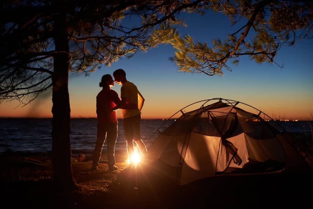 Silhouettes of hiker couple, back view of a man and woman standing at a campfire embracing near a tourist tent in evening.