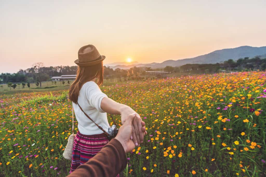 A woman leads a man into a field of flowers at sunset.