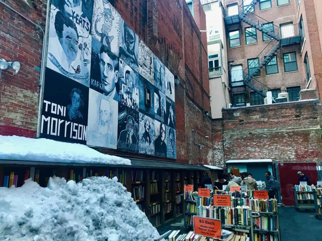 Blue and grey street art on the side of a brick building that says \"Toni Morrison\" next to an outdoor book shop.