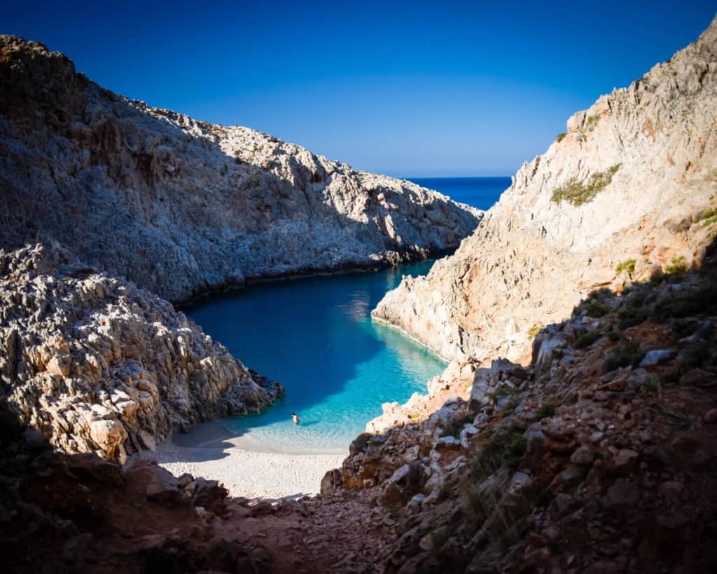 A small strip of water between two rocky cliffs with a small sandy beach at the bottom. 