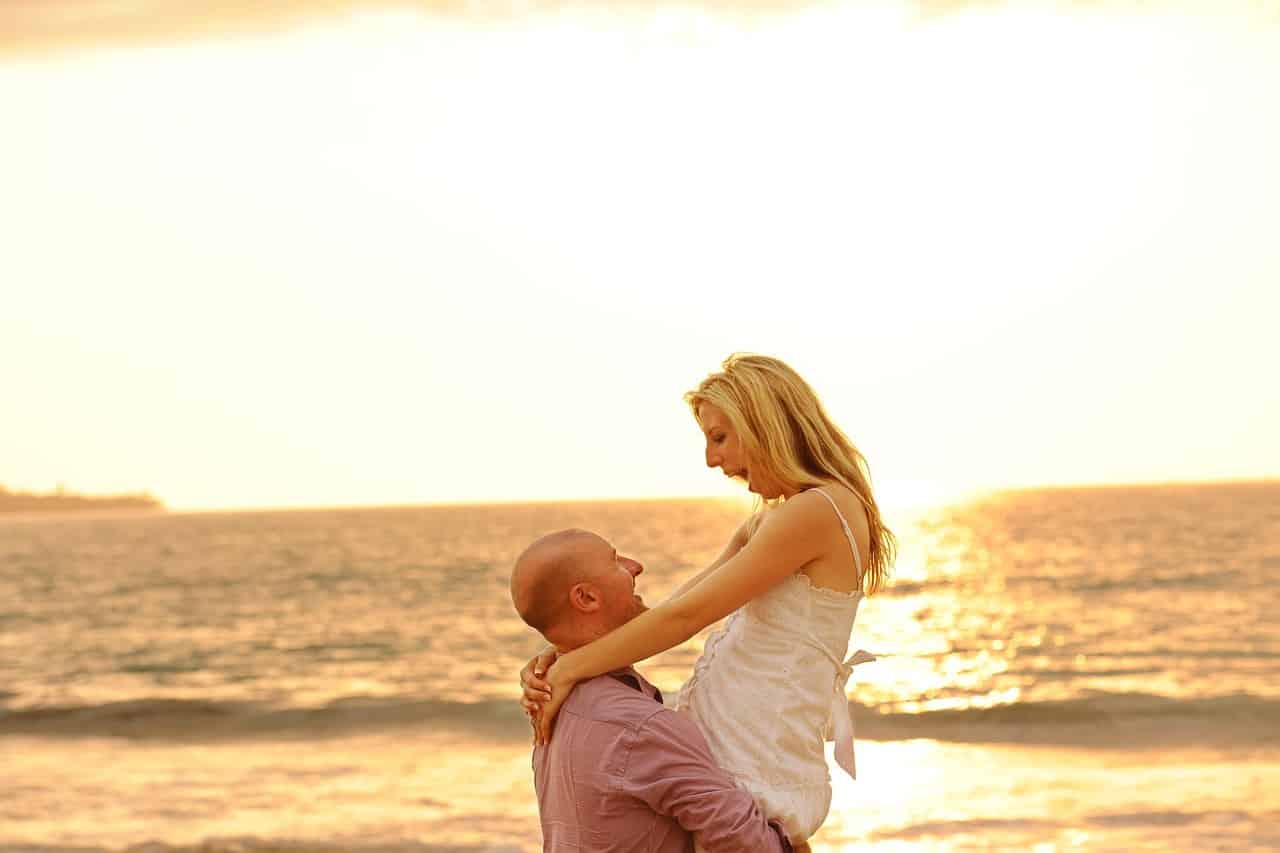 A man picks up a woman in front of the ocean.