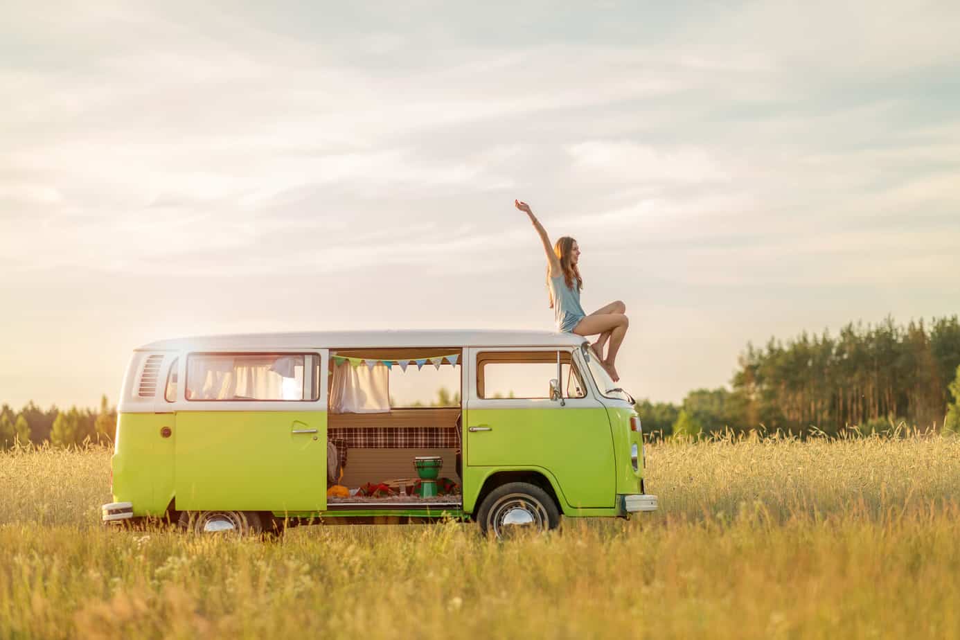 A woman sits on top of a green van with her arms in the air in the middle of a field.
